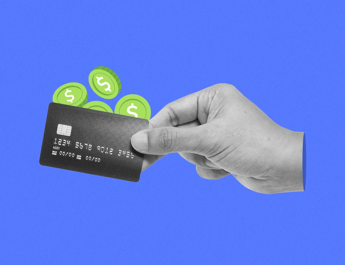 Una tarjeta de crédito siendo sostenida por una mano mientras varias monedas verdes están detrás de ella.
