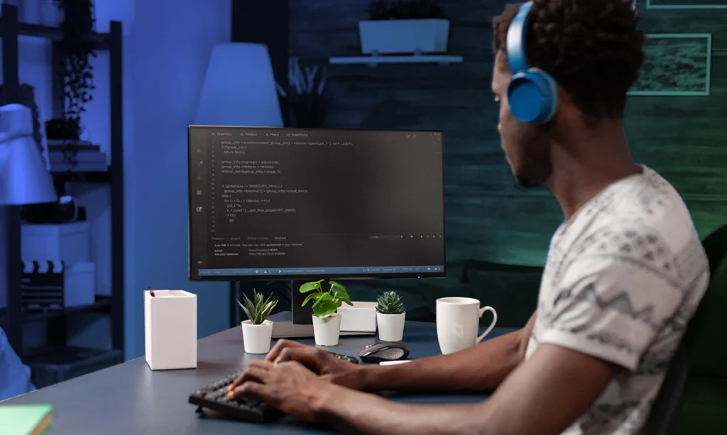 A young man focused on coding on his computer in a home office setup, he is wearing over-the-ear headphones.